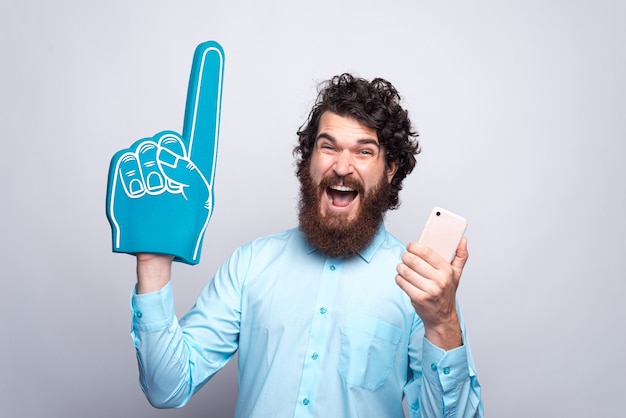 Foto des verblüfften bärtigen Mannes im blauen Hemd, das Telefon und Fan Foam Glove hält