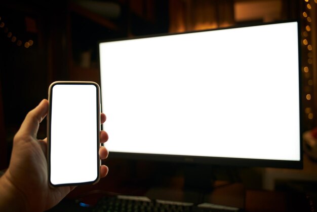 Foto des weißen leeren Bildschirms des Computers und der Hand, die das Mobiltelefon hält Digitales Webspace-Konzept