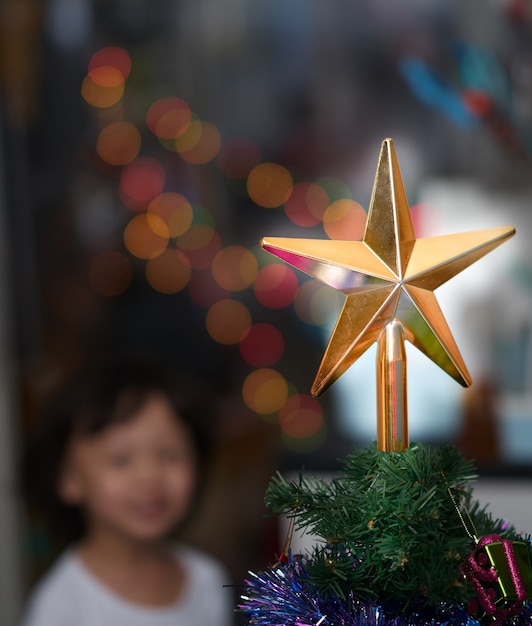 Foto de desenfoque de sonrisa de niño mientras mira la decoración de estrella de oro en el árbol de Navidad con bokeh