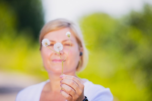 Foto desfocada de uma mulher loira ao ar livre, segurando a flor dente de leão na mão e vestindo roupa elegante.