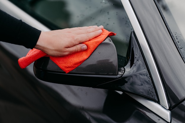 Foto detalhada da mão do proprietário limpando o espelho lateral do carro preto com um pano de microfibra. Conceito de limpeza e cuidado com o carro