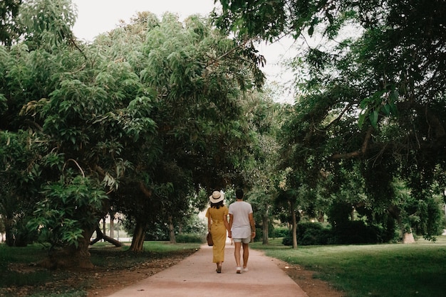 Una foto por detrás de una chica morena con un vestido amarillo y su novio que caminan por un camino de arena entre árboles en el parque valenciano. Un par de turistas en una cita en la cálida noche.