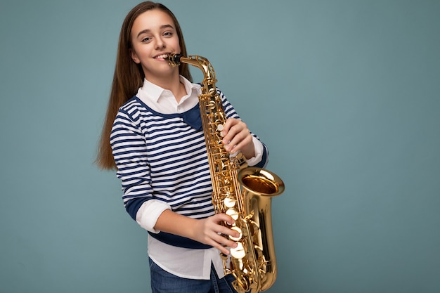 Foto disparada de uma linda menina morena sorridente feliz vestindo pé de manga comprida listrado isolado sobre a parede de fundo azul tocando saxofone, olhando para a câmera. Copie o espaço