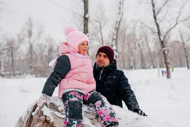 Foto distante de una niña sentada en un tronco de árbol sonriendo y mirando hacia otro lado usando ropa rosa de invierno...