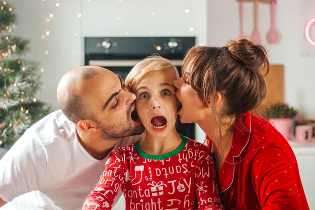 Foto divertida de la familia en pijama de Navidad en la cocina muerde alegremente las mejillas de los niños