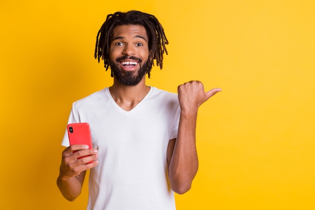 Foto de divertido asombrado chico de piel oscura mantenga el teléfono dirigiendo el dedo espacio vacío novedad aislado de fondo de color amarillo