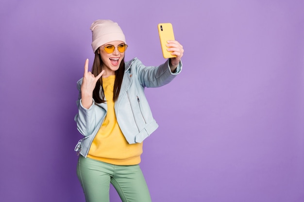 Foto de divertido bastante guay joven dama sostener teléfono hacer selfies blogger popular mostrando cuernos de dedo usar especificaciones de sol chaqueta de sombrero casual pantalones de jersey amarillo fondo de color púrpura aislado