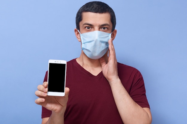 Foto do cara chocado emocional, colocando uma mão perto do rosto, segurando o smartphone, entrando em pânico, usando o dispositivo, ficando em casa, assustada pelo coronavírus. pessoas e conceito de pandemia.