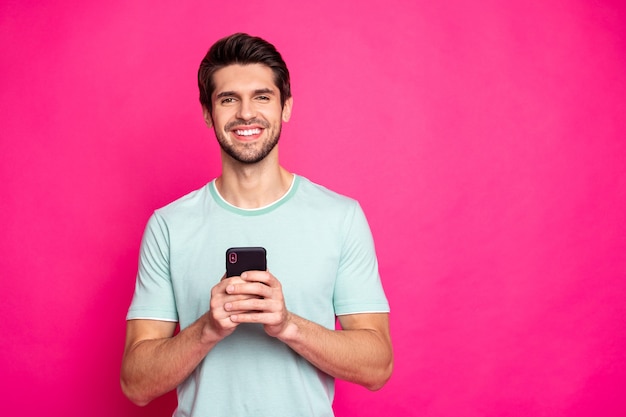 Foto do cara do blogueiro segurando o telefone nas mãos, verificando os assinantes, sorrindo, vestindo roupa casual isolado fundo de cor rosa viva