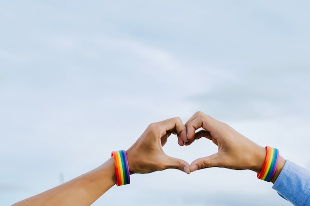 foto do close up de um casal gay de mãos dadas com uma pulseira de arco-íris formando um coração