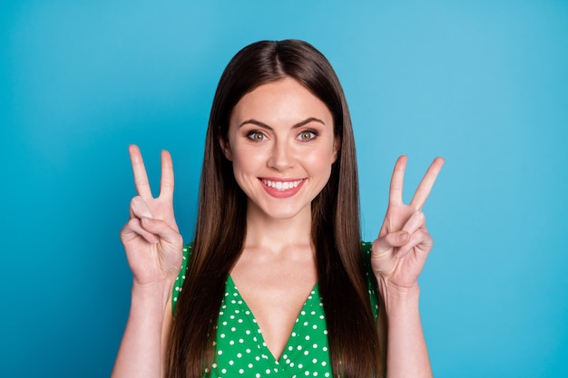 Foto do close up de uma senhora atraente, bom humor, sorriso branco e encantador, aparência fofa mostrando os símbolos do sinal de V, as mãos vestem uma camiseta casual verde pontilhada isolada no fundo da cor azul