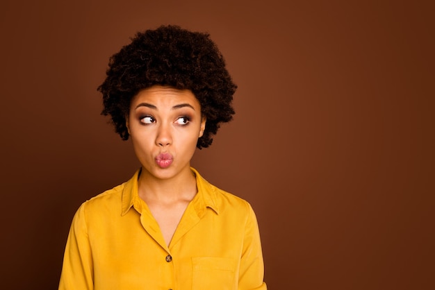 Foto do close up de uma senhora encaracolada de pele muito morena bonita parecendo tímido lado vazio evite olhos de cara bonito enviando-lhe beijo no ar usar camisa amarela isolada cor marrom