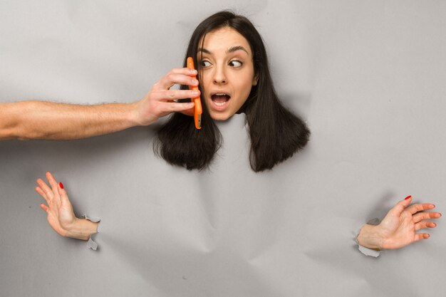 Foto do conceito de jovem fêmea rasgando papel e espreitando o buraco, a mão do homem dando smartphone, fundo de cor cinza isolado.