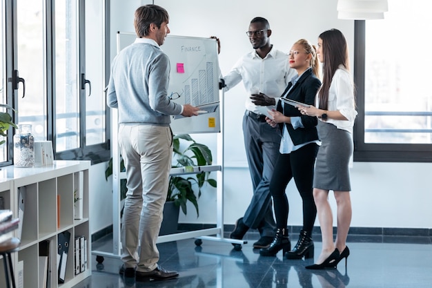 Foto do empresário bonitão apontando para a lousa enquanto explica o projeto para seus colegas no local de coworking.