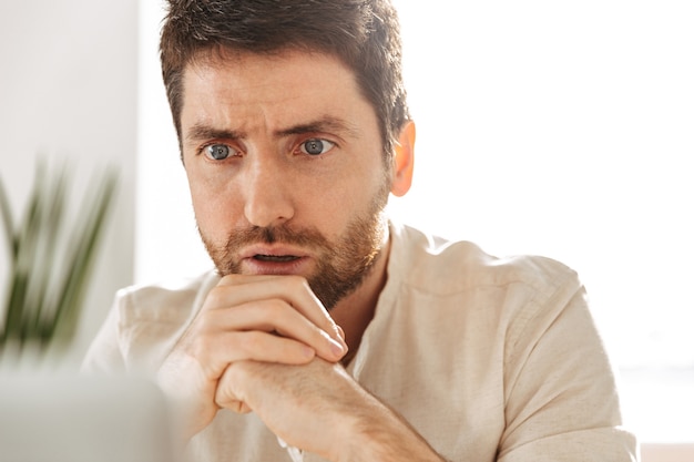 Foto do empresário confiante dos anos 30, vestindo uma camisa branca, usando um laptop, enquanto está sentado à mesa em um escritório moderno