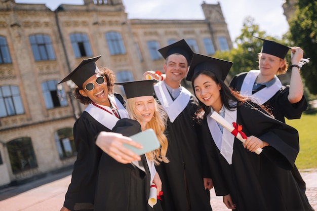 Foto do grupo. colegas de grupo cherful em frente à universidade tirando selfies após a formatura.