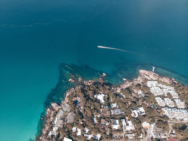 Foto do helicóptero. Pequena cidade estrangeira perto da praia de big kata. Barcos flutuam na água. Toda a cidade está coberta de árvores verdes e palmeiras. Muitos edifícios e locais diferentes.