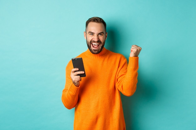 Foto do homem feliz triunfando, segurando o telefone celular e fazer o gesto do vencedor, alegrar-se e alcançar o objetivo, de pé sobre o fundo azul claro.