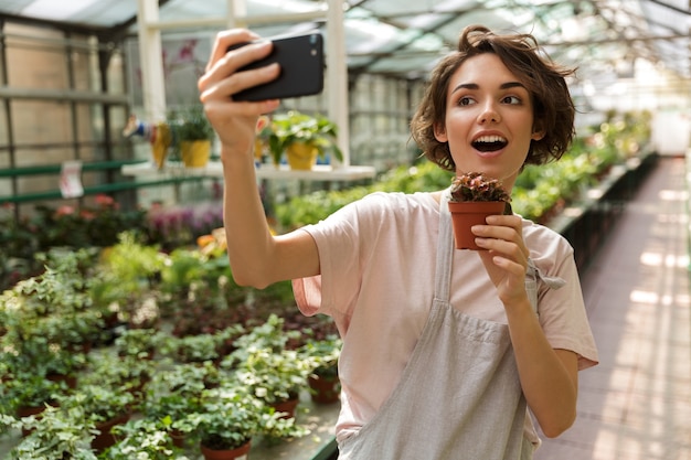 Foto do jardineiro de mulher bonita bonita em pé sobre plantas de flores em estufa faz selfie pelo telefone celular.