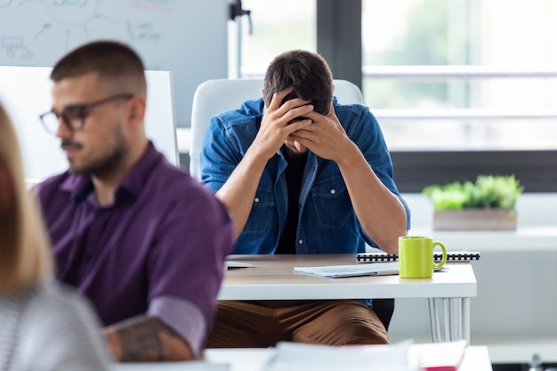 Foto do jovem empresário segurando seu rosto com as mãos enquanto está sentado à mesa no escritório criativo. Dia estressante no escritório.