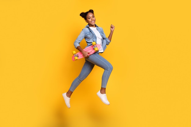 Foto do lado do perfil de corpo inteiro de uma jovem africana de bom humor, vá caminhar, corra, segure o skate isolado sobre o fundo amarelo