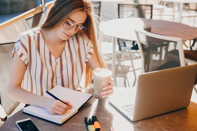 Foto do lado superior de uma garota legal que está trabalhando em um caderno, sentada em frente ao computador com uma xícara de café