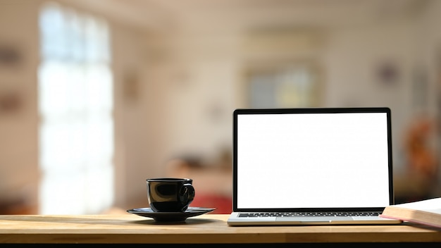 Foto do laptop de tela em branco branco, xícara de café preto e montanhas-russas na mesa de trabalho de madeira sobre café moderno turva