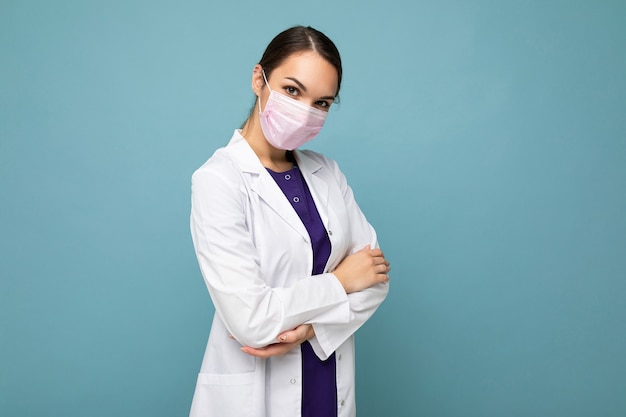 Foto do médico da bela jovem de jaleco branco e máscara médica em pé isolado sobre o fundo azul. Conceito de coronavírus