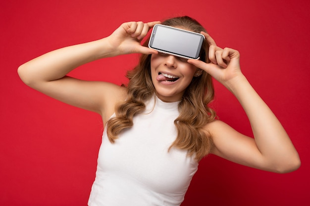 Foto do retrato de uma jovem loira engraçada vestindo uma camiseta branca isolada em um fundo vermelho com espaço de cópia segurando o smartphone mostrando o telefone na mão com uma tela vazia para recorte e mostrando a língua.