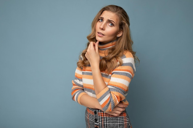 Foto do retrato de uma jovem muito bonita atraente triste mulher loira infeliz com cabelos ondulados, vestindo uma camisola listrada isolada sobre um fundo azul com espaço vazio para o texto.