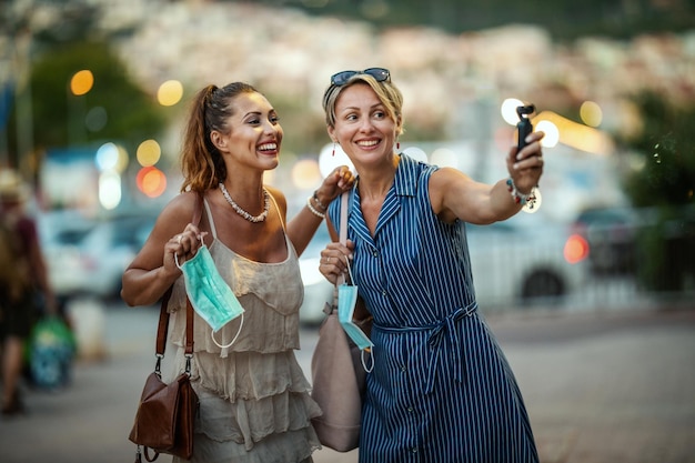 Foto de dos amigas atractivas que usan una máscara quirúrgica y usan una cámara digital mientras exploran la maravillosa ciudad de Meditteranian.