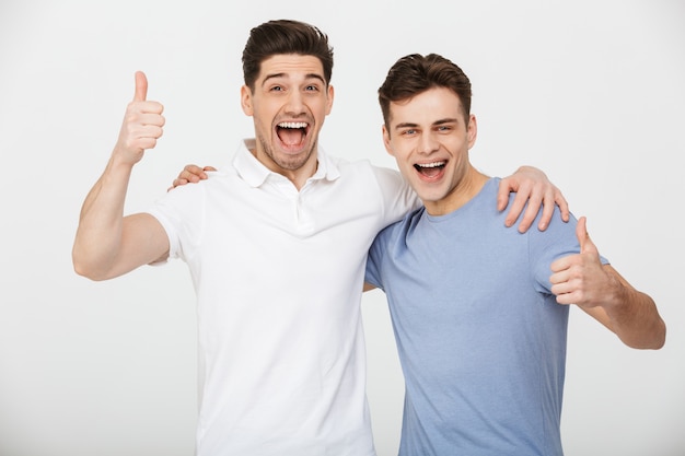 Foto foto de dos amigos varones 30s vistiendo camiseta casual y jeans sonriendo y mostrando los pulgares hacia arriba en la cámara, aislado sobre fondo blanco.