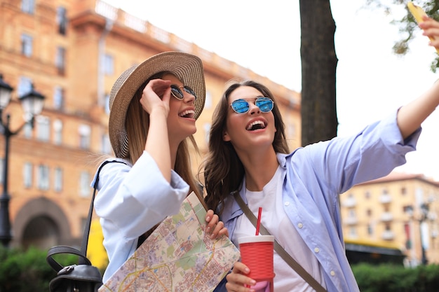 Foto de dos chicas disfrutando de hacer turismo al aire libre. Hermosas mujeres turistas explorando la ciudad con mapa y tomando selfie.