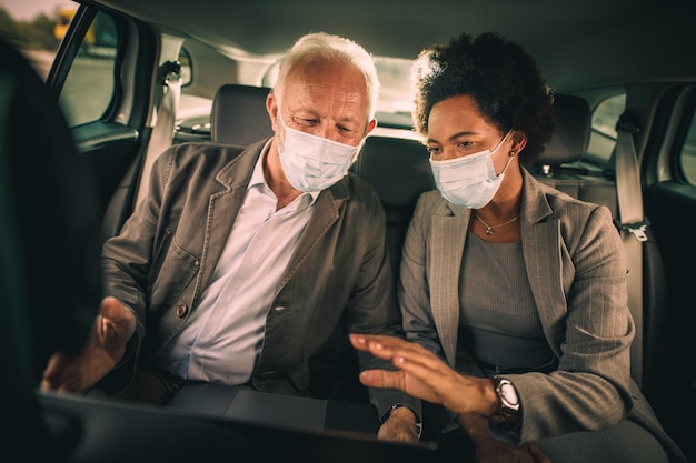 Foto de dos exitosos empresarios multiétnicos con máscara protectora usando una laptop y discutiendo mientras estaban sentados en el asiento trasero de un camión en su viaje matutino durante la pandemia de COVID-19.