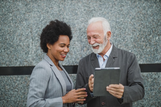 Foto de dos exitosos empresarios multiétnicos que usan una tableta digital y tienen una discusión mientras están de pie contra una pared de un edificio corporativo.