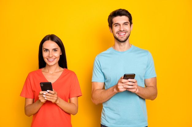 Foto de dos graciosos chico dama gente pareja sostener teléfonos brazos leer comentarios de publicaciones de redes sociales usar camisetas casuales azules naranjas pared de color amarillo aislado