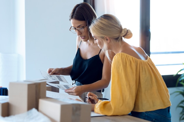 Foto de dos hermosas mujeres de negocios independientes que comprueban el pedido del producto mientras empacan y sellan cajas de cartón para los productos de entrega pedidos en línea a los clientes en su pequeña empresa de inicio