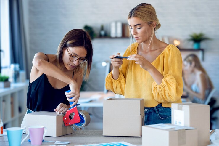 Foto foto de dos hermosas mujeres de negocios independientes que toman fotos con un teléfono inteligente mientras empacan y sellan cajas de cartón para la entrega de productos pedidos en línea a los clientes en su pequeña empresa de inicio