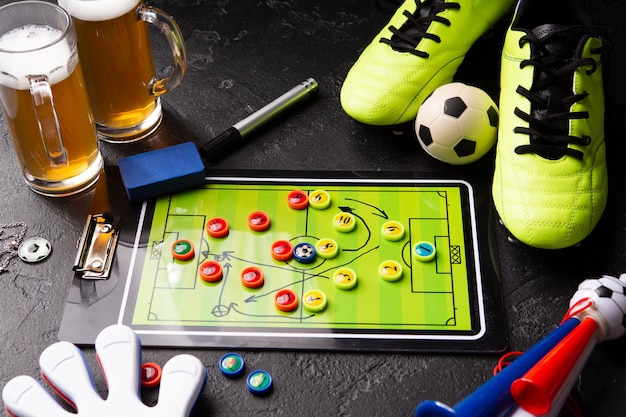 Foto de dos jarras de cerveza de espuma, futbolín, pelota, botas de fútbol, pipa, sonajero en mesa negra