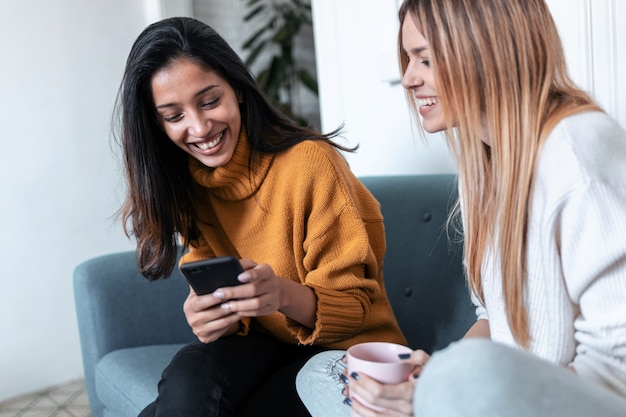 Foto de dos mujeres bastante jóvenes con teléfono móvil mientras bebe café en el sofá en casa.