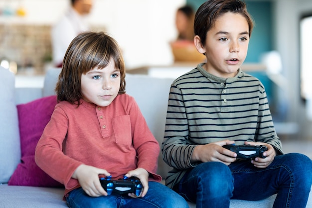 Foto de dos simpáticos hermanos jugando videojuegos mientras está sentado en el sofá de casa.