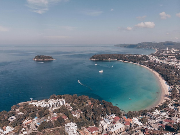 Foto de drones de una ciudad turística atractiva y atmosférica con varios edificios, árboles y palmeras. Detrás hay un enorme océano claro con una playa de arena kata en la costa. En el centro de la mini isla.