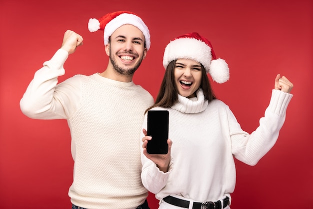 Foto de dulce pareja con sombreros de Navidad, tiene nuevo teléfono inteligente para regalo. Hombres y mujeres están enamorados, se ven bendecidos y sonríen, aislados sobre fondo rojo.