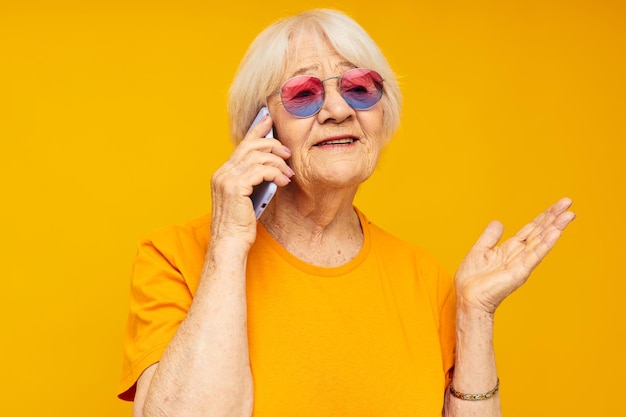 Foto einer alten Dame im Ruhestand, die am Telefon in gelber Brille spricht, gelber Hintergrund