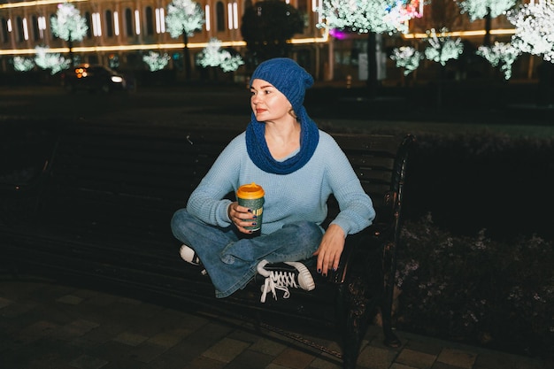 Foto einer attraktiven, fröhlichen Frau, die bei kaltem Schneewetter auf einer Bank sitzt, lächelt und draußen Kaffee trinkt