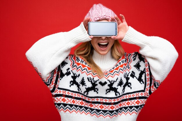 Foto einer attraktiven lächelnden jungen blonden Frau mit warmer Strickmütze und warmem Winterpullover, die isoliert auf rotem Hintergrund steht und Smartphone mit leerem Bildschirm für den Ausschnitt zeigt