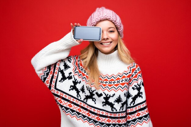 Foto einer attraktiven lächelnden jungen blonden Frau mit warmer Strickmütze und warmem Winterpullover