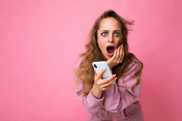 Foto einer attraktiven, verrückten überraschten jungen Frau, die lässige, stilvolle Kleidung trägt, die isoliert über dem Hintergrund steht, mit Kopienraum, der das Mobiltelefon hält und in die Kamera schaut.