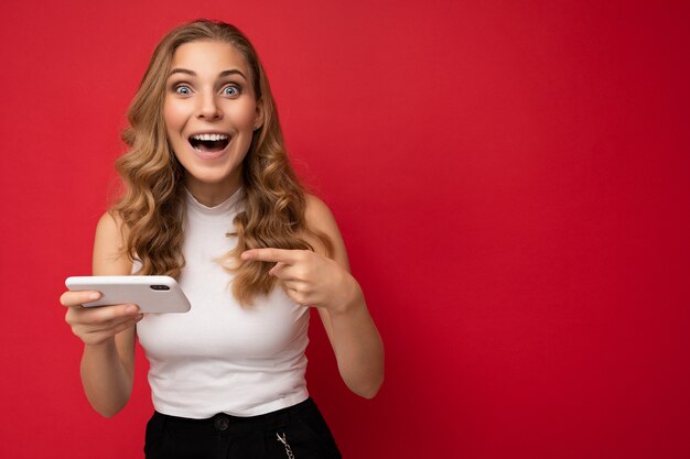 Foto einer attraktiven, verrückten überraschten jungen Frau, die lässige, stilvolle Kleidung trägt, die isoliert über dem Hintergrund steht, mit Kopienraum, der das Mobiltelefon hält und in die Kamera schaut