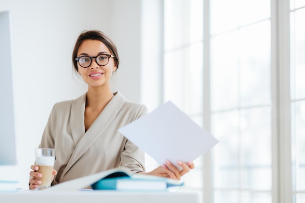 Foto einer entzückten dunkelhaarigen Europäerin mit transparenter Brille arbeitet mit Dokumenten liest Informationsdaten in eleganter Kleidung trinkt Milchshake sitzt am Schreibtisch im Büro macht Bericht